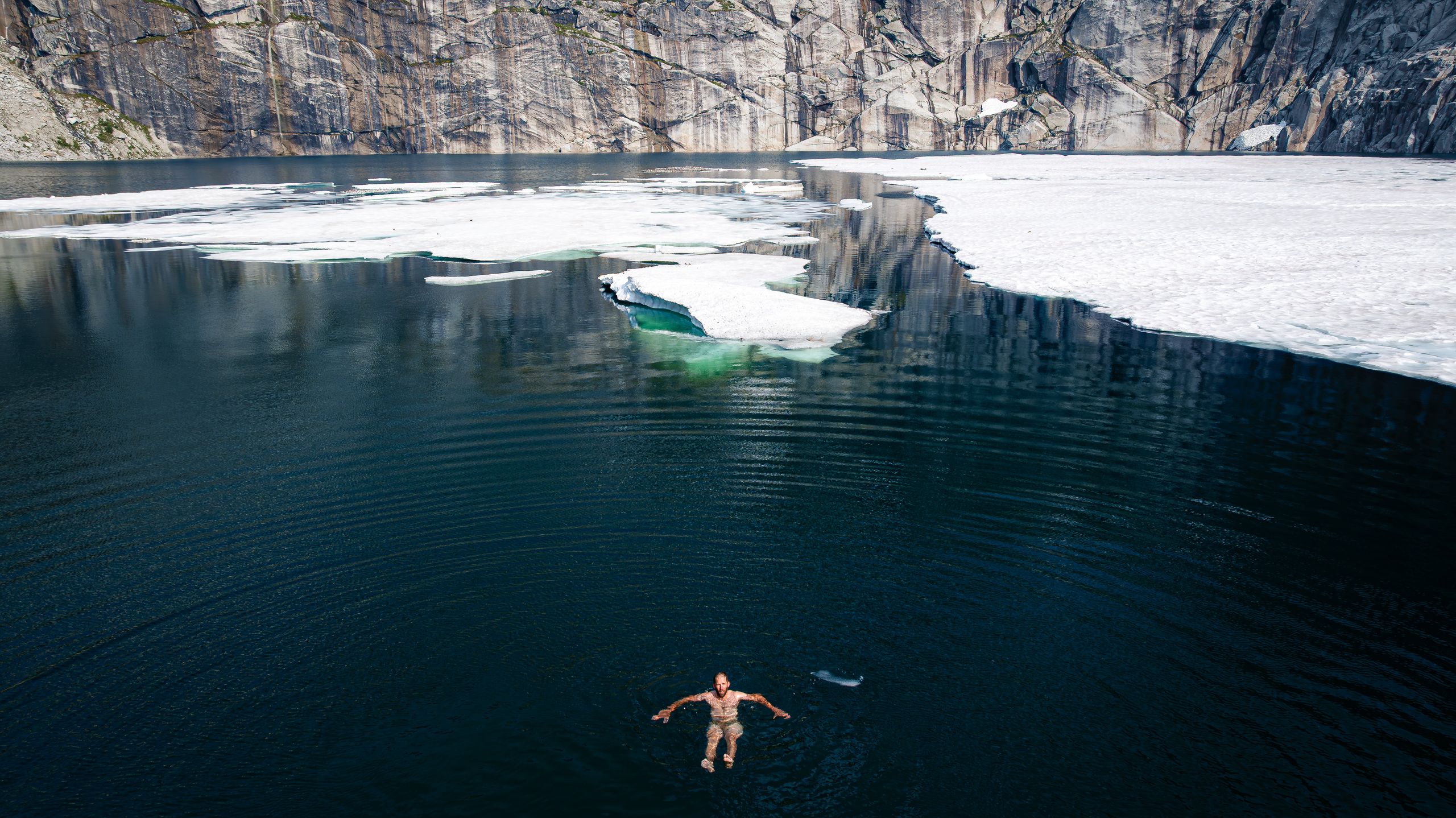 A swimmer floating in a dark alpine lake edged with ice