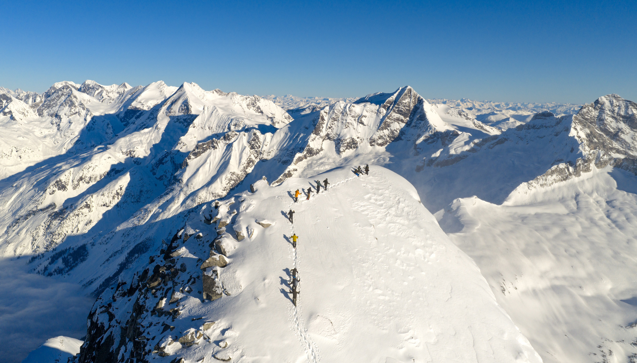 Ski tourers in single file on a summit ridge, with the Selkirks all around