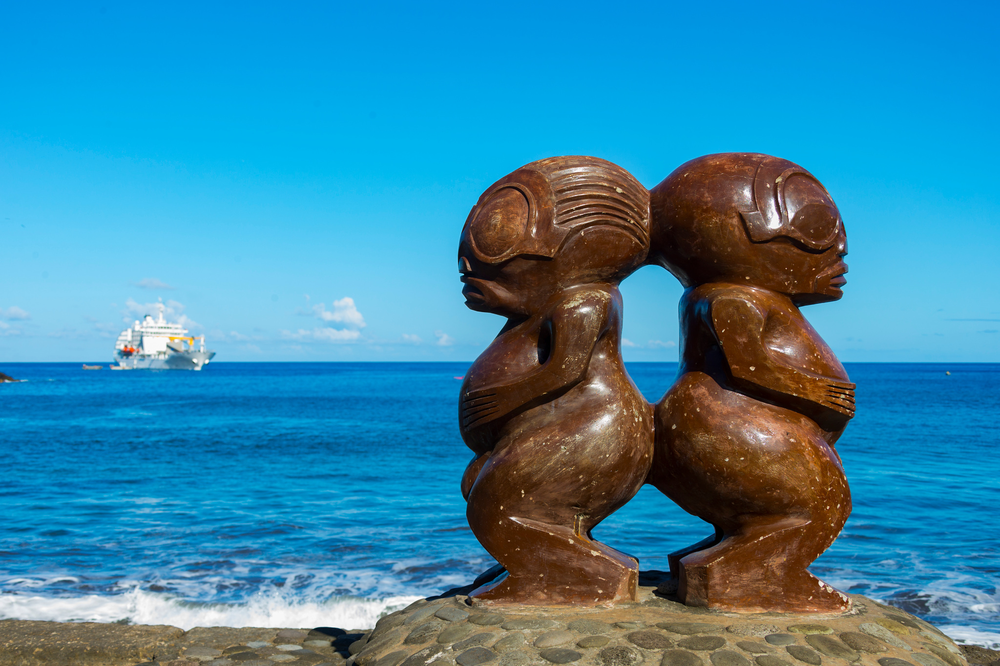Two carved wooden tikis back to back on a seawall, Aranui 5 offshore