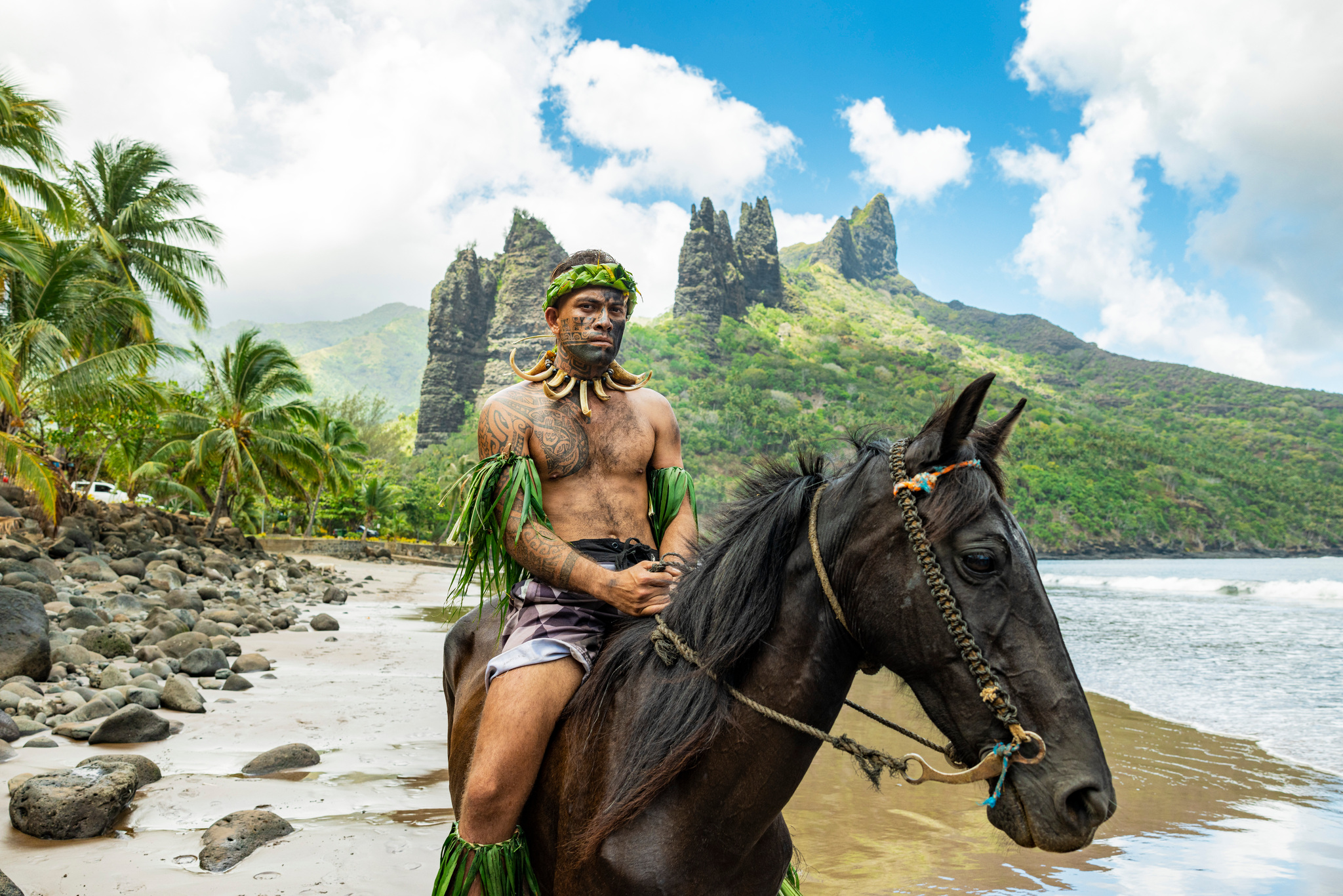 A tattooed rider in a leaf headdress on a dark horse on the beach at Hatiheu, rock spires behind