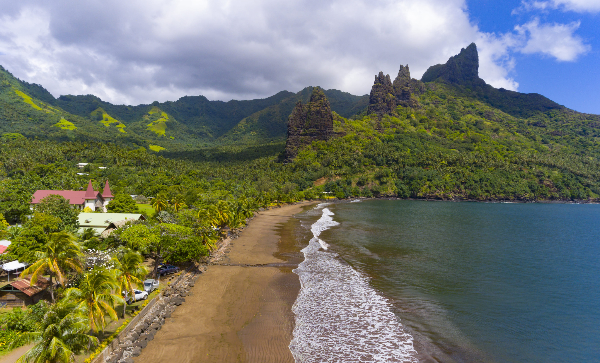 Hatiheu village and its church beside a curving black-sand bay below green peaks on Nuku Hiva, from the air