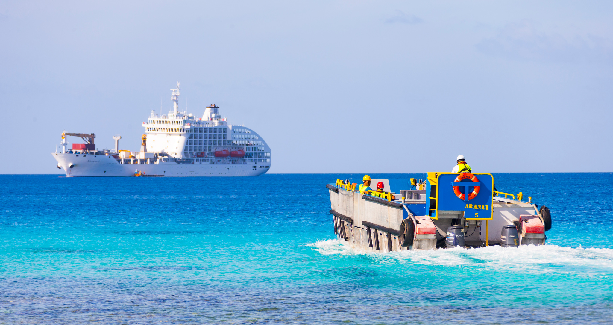 The landing barge and its crew crossing a turquoise lagoon, Aranui 5 at anchor behind