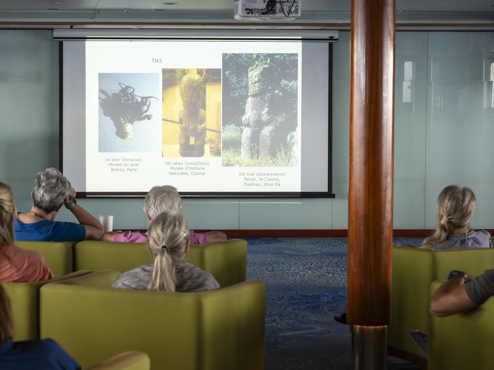 Passengers in armchairs at a lecture, with photographs of tikis on the screen