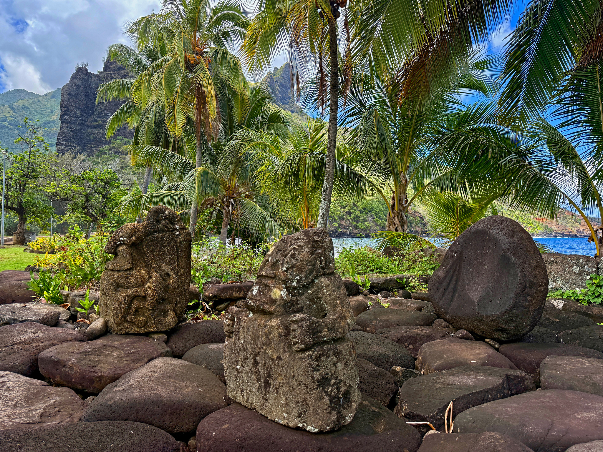 Stone tikis on a paved platform among palms, below the peaks of Nuku Hiva