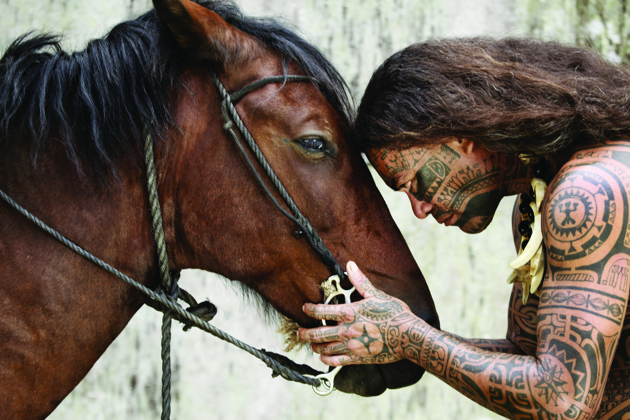 A tattooed Marquesan man resting his forehead against his horse's