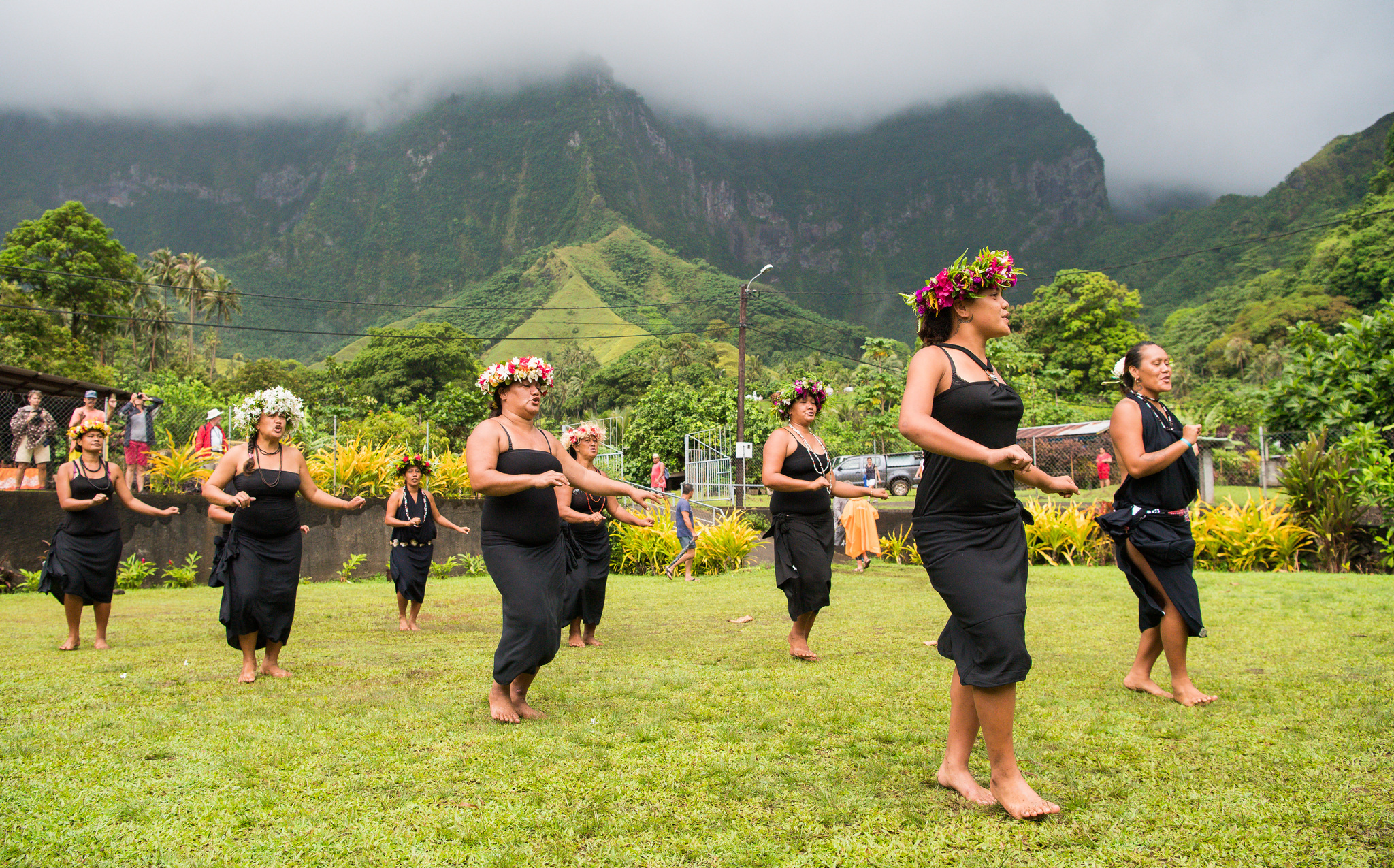 Women in black dresses and flower crowns dancing on a village green below misty mountains