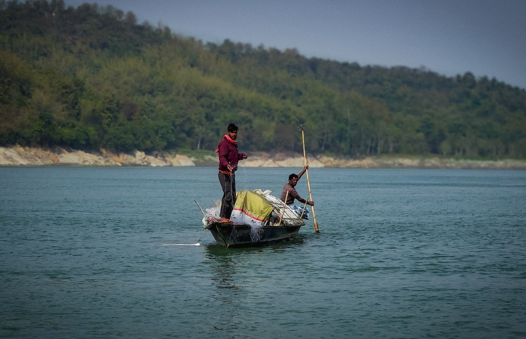 A boatman poling a loaded boat below a forested hill
