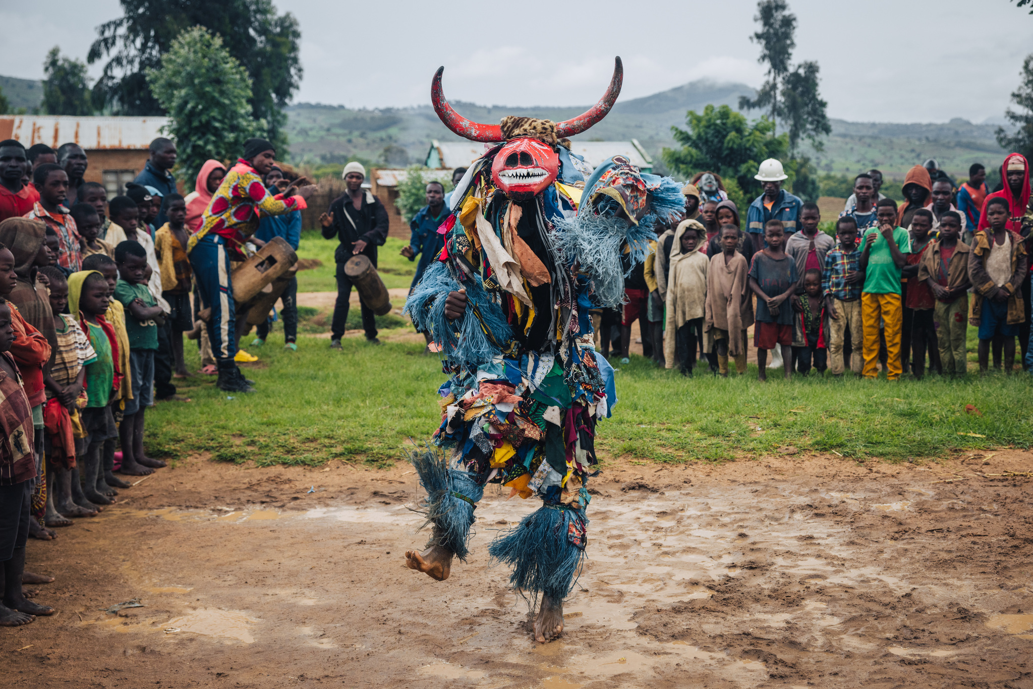 A Gule Wamkulu dancer in a horned mask and a costume of rags, mid-leap before the village