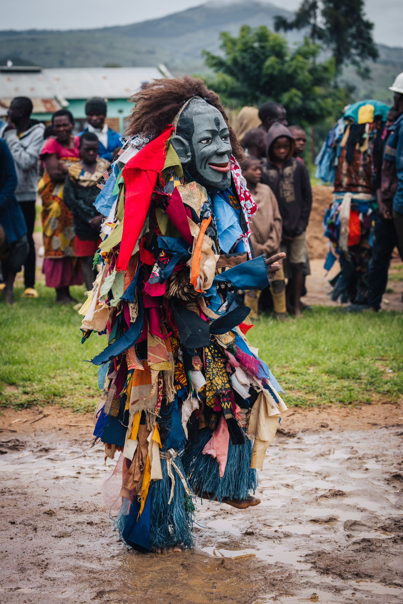 A dancer in a pale mask and a costume of coloured rags
