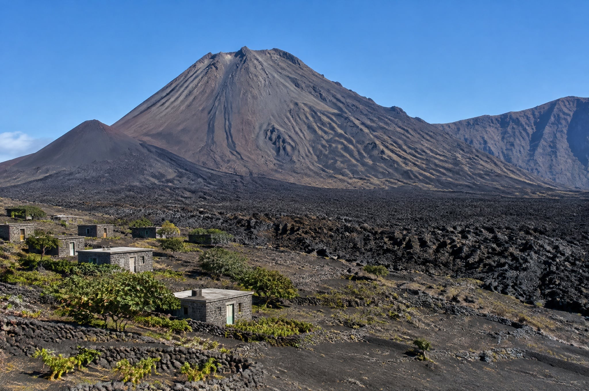 Pico do Fogo rising behind stone houses, vines and black lava fields at Cha das Caldeiras, inside the Fogo caldera, Cabo Verde