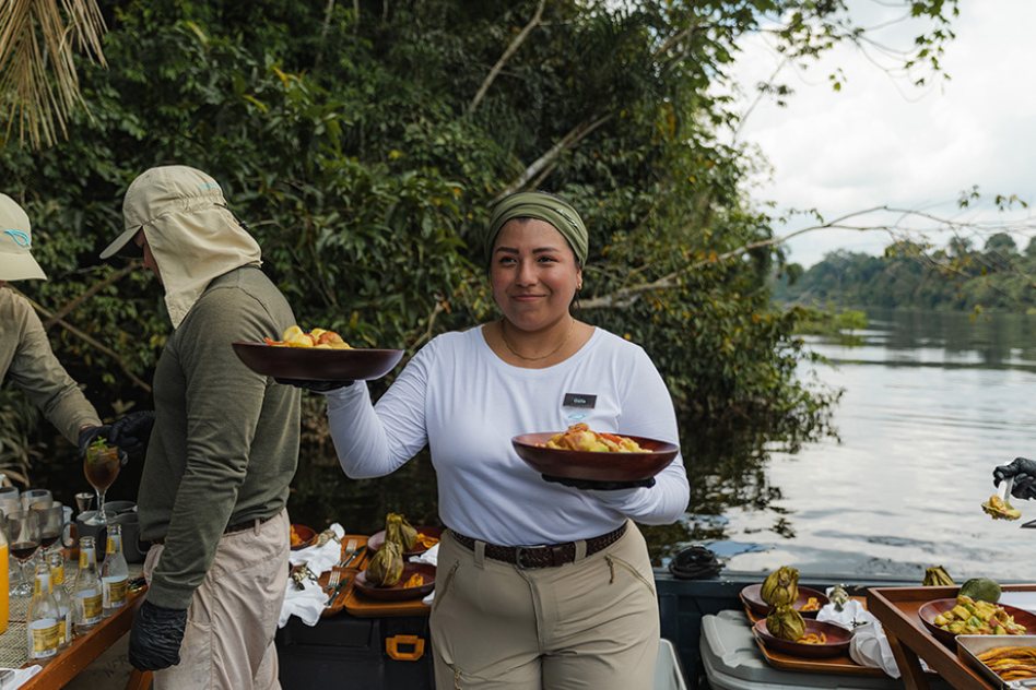 A crew member serving breakfast at a table set up in the forest by the river