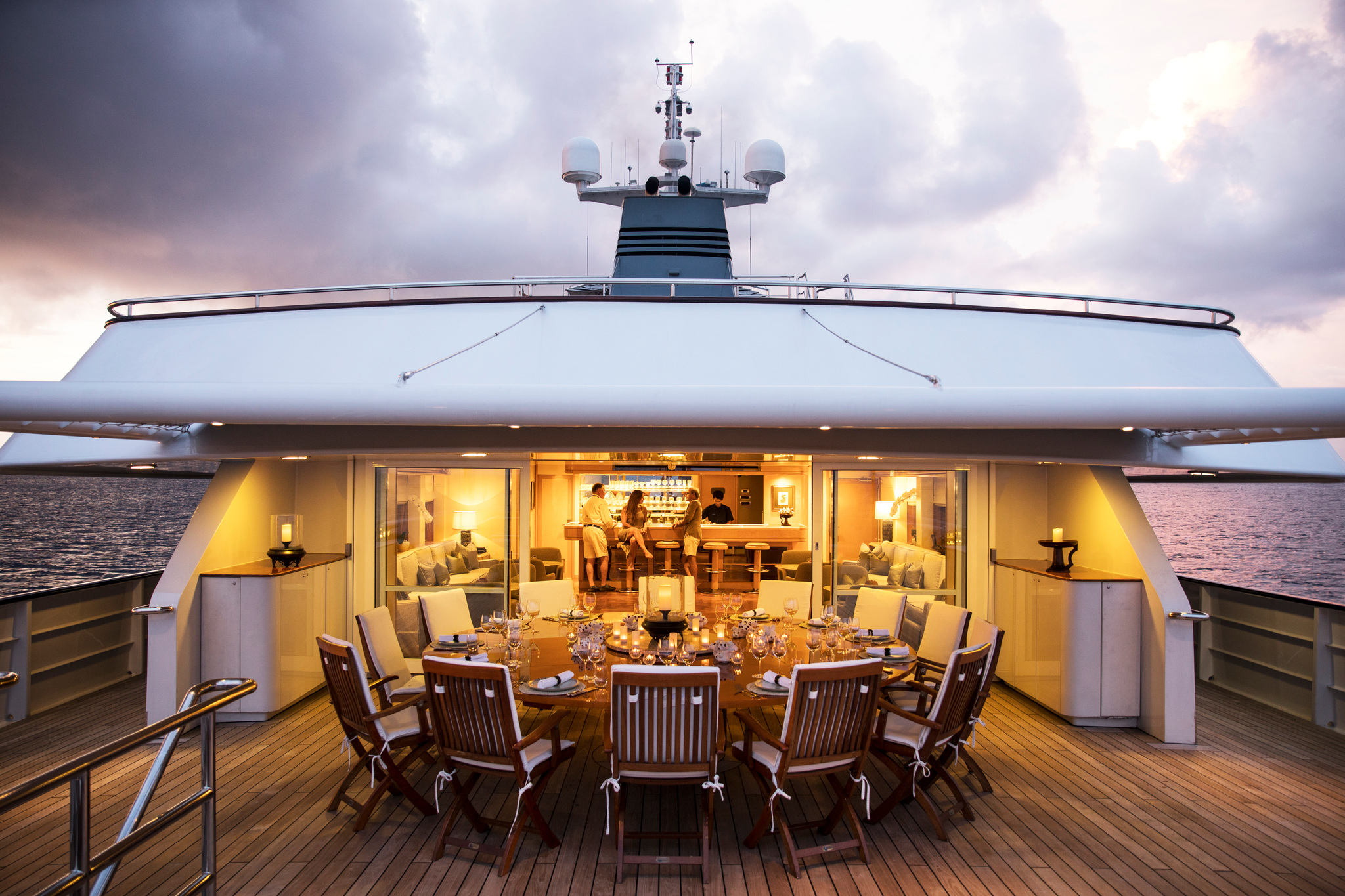 A round table laid for dinner on the open deck at dusk, in front of the lit lounge