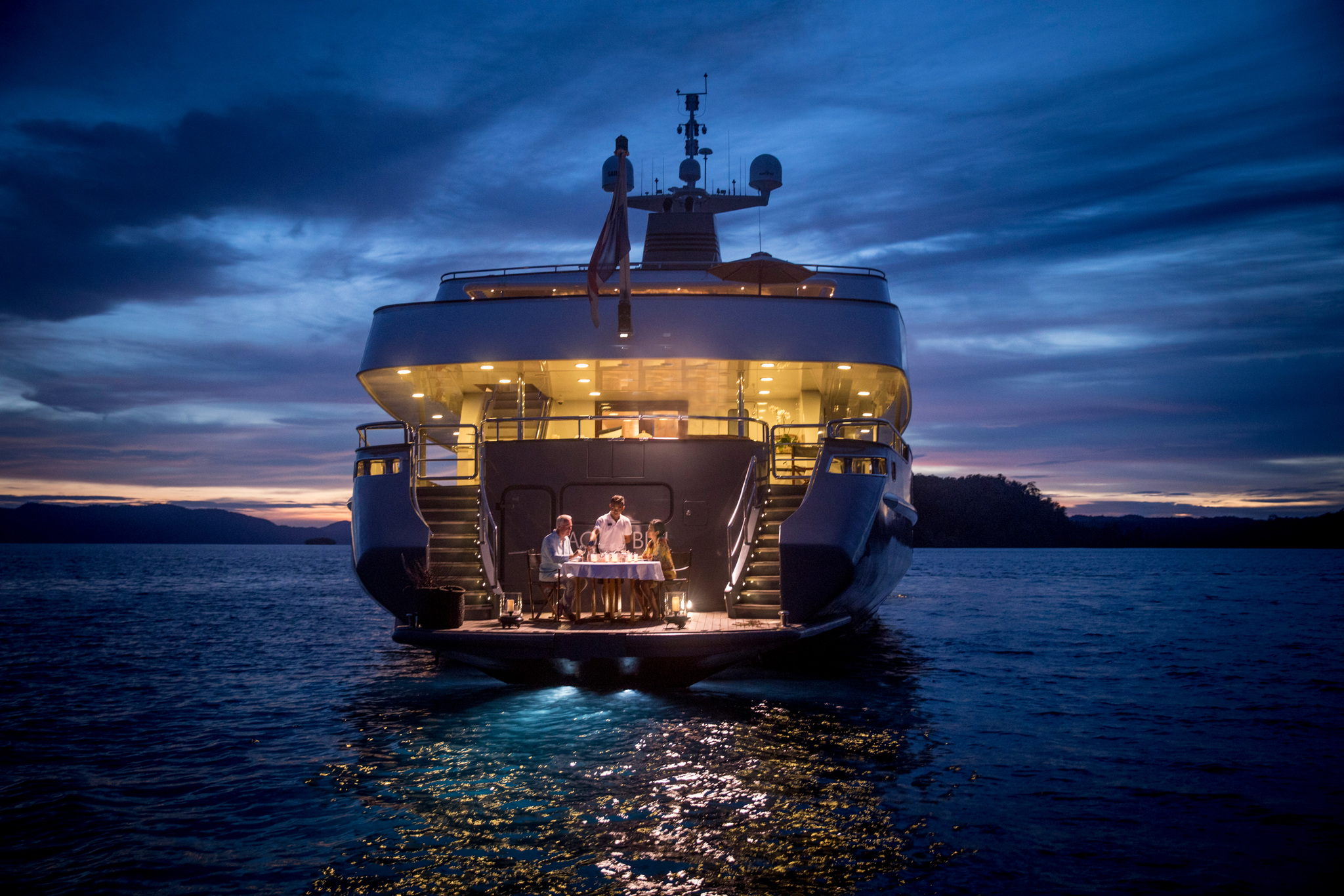 Two guests dining at a candlelit table on the stern platform at dusk, the decks lit above