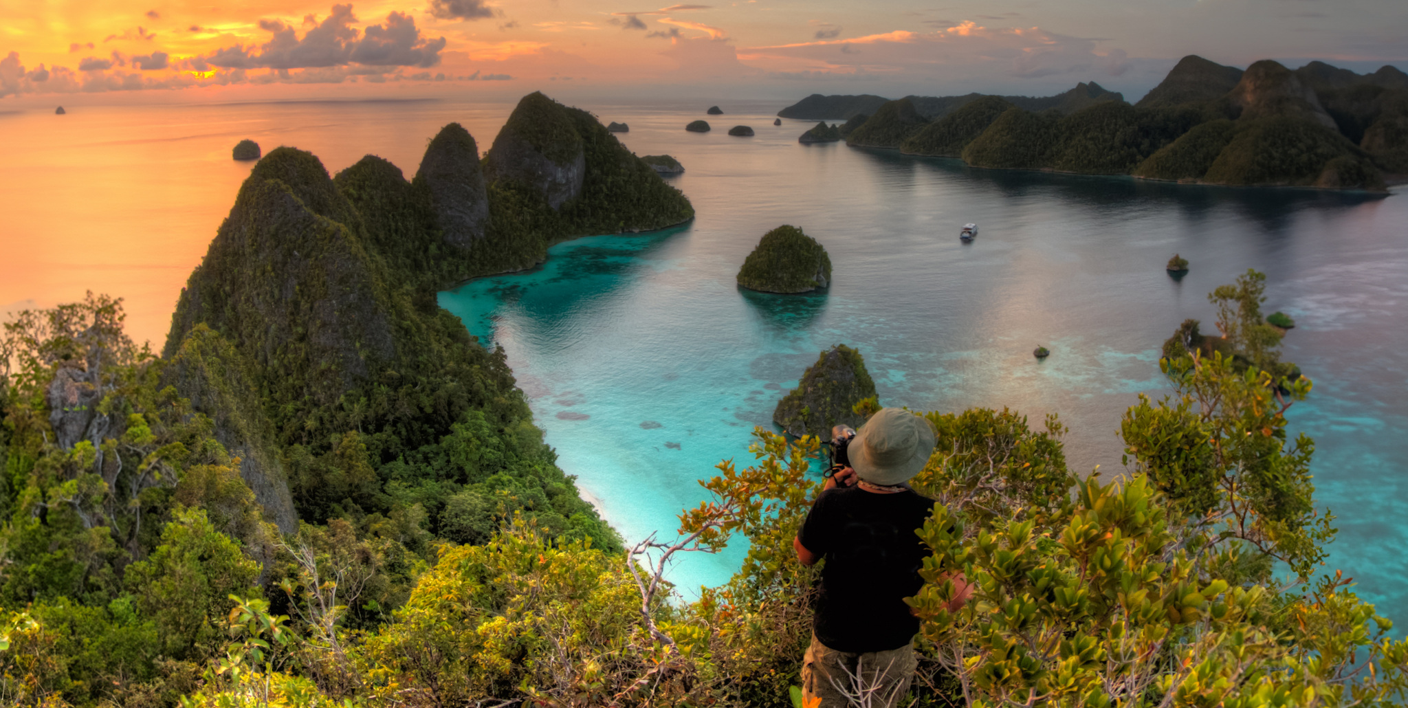 A guest on a hilltop photographing forested karst islands and turquoise channels at sunset