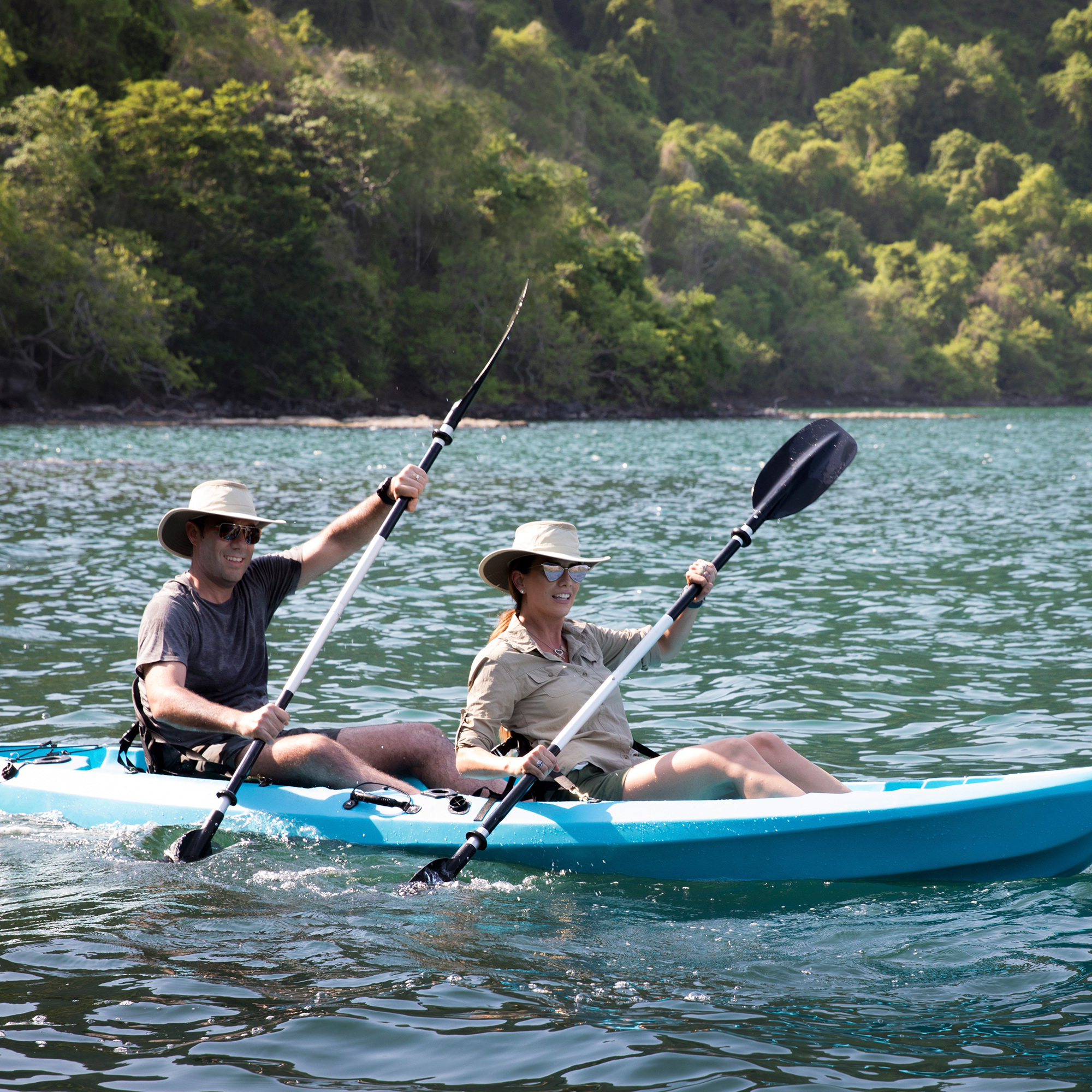 Two people in sun hats paddling a double kayak past a forested shore