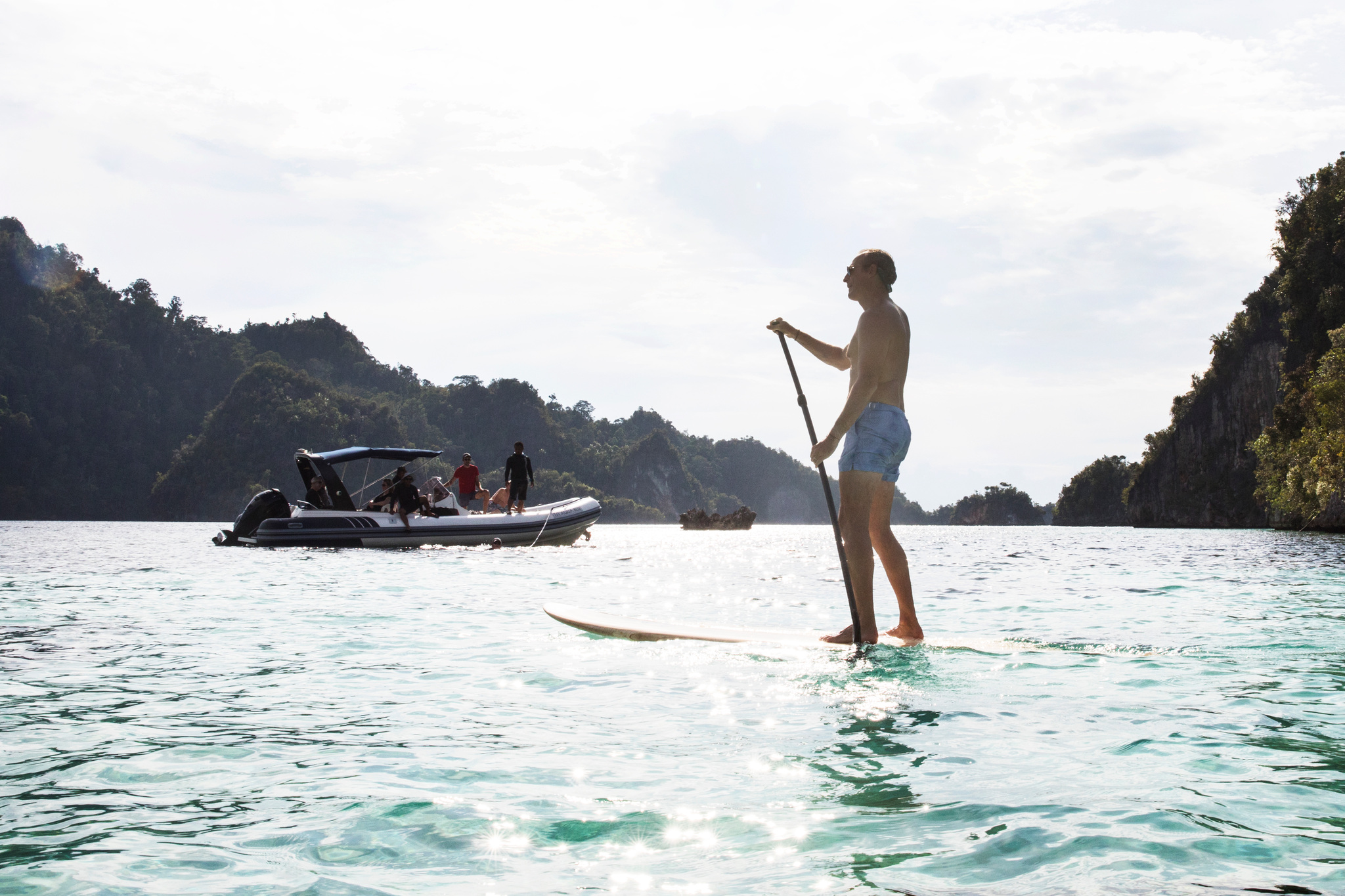 A man on a paddleboard in clear shallows between limestone cliffs, a tender behind