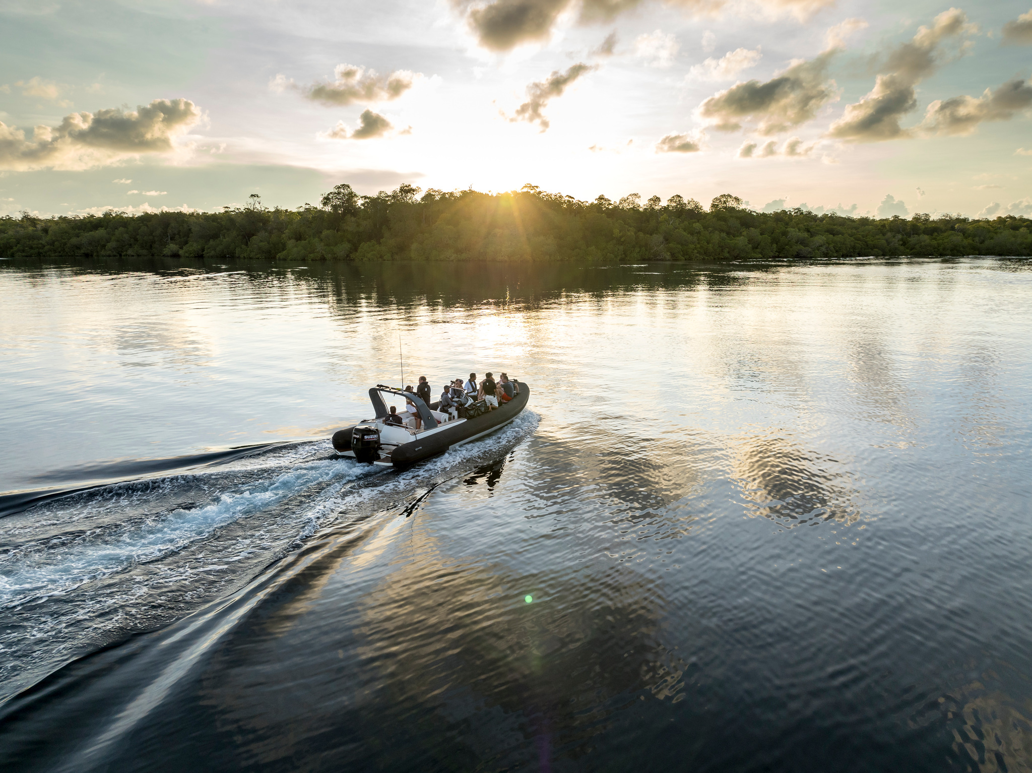 A tender of guests crossing glassy water toward a forested shore, the sun low over the trees